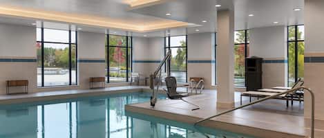 Una piscina interior con paredes de vidrio transparente, una escalera de piscina y una zona de estar con sillas y mesas.