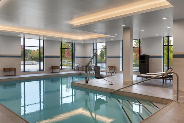 Una piscina interior con paredes de vidrio transparente, una escalera de piscina y una zona de estar con sillas y mesas.
