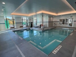 Een groot binnenzwembad met helder water, omringd door betegelde vloeren en wanden. Er zijn banken en een kleedruimte te zien.