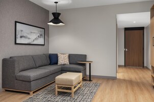 Una sala de estar moderna con un sofá seccional gris, un pequeño banquillo de madera, una mesita redonda, una lámpara colgante negra y un cuadro abstracto enmarcado en la pared.