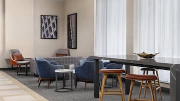 Una habitación de hotel moderna con zona de comedor, sillas tapizadas en azul, un sofá gris y un ventanal con cortinas translúcidas.