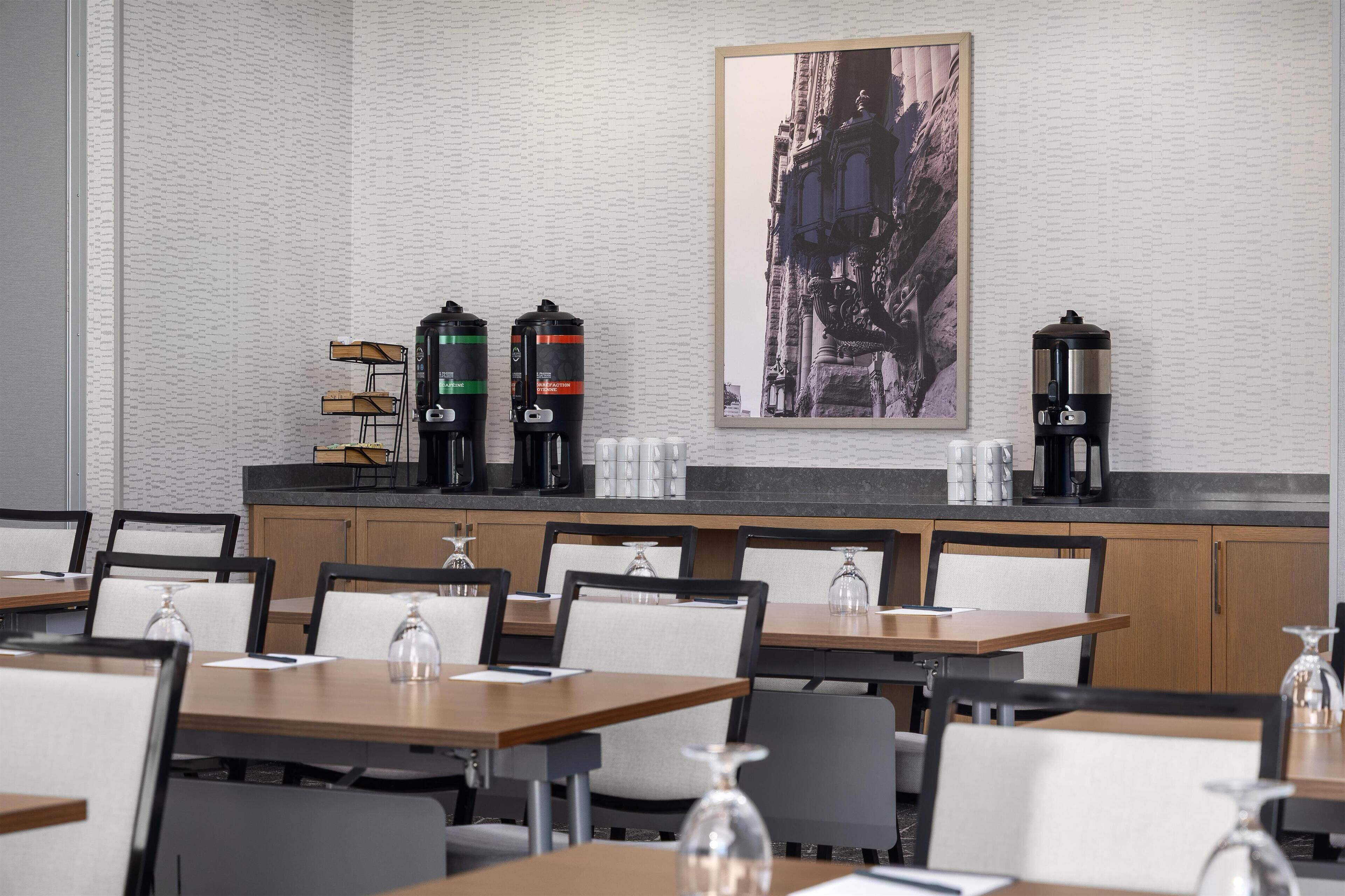 A modern hotel dining area with multiple tables and chairs, a coffee station with three coffee makers, and a framed cityscape artwork on the wall.