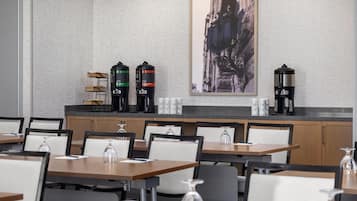 A modern hotel dining area with multiple tables and chairs, a coffee station with three coffee makers, and a framed cityscape artwork on the wall.