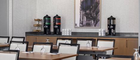 A modern hotel dining area with multiple tables and chairs, a coffee station with three coffee makers, and a framed cityscape artwork on the wall.