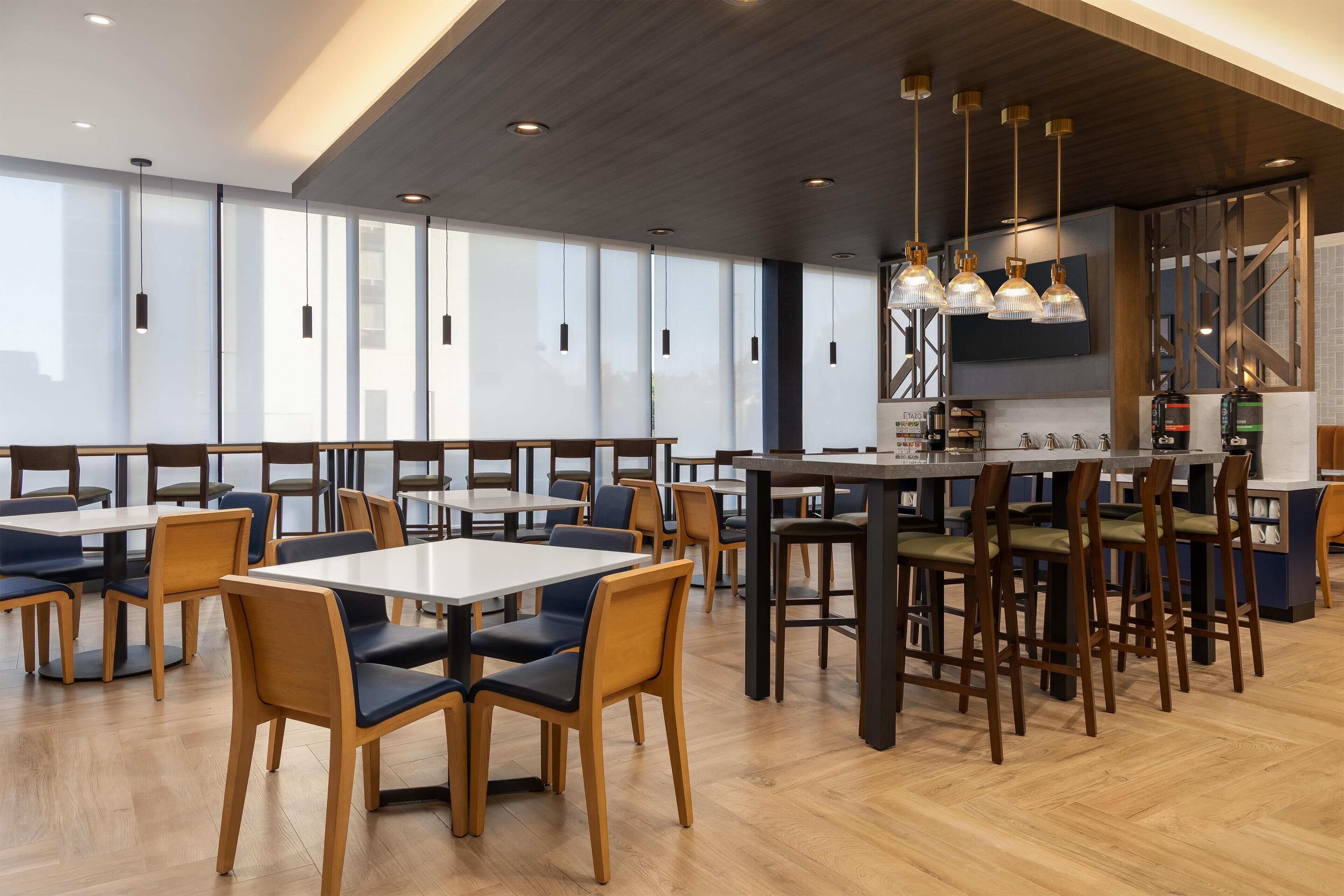 A modern dining area with wooden tables and chairs, pendant lighting, and a kitchen area with appliances.