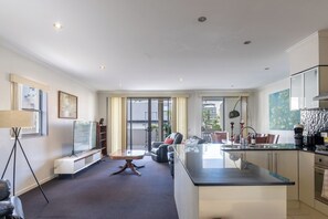 A modern hotel room with a large central island, a seating area with a sofa and chairs, a flat-screen TV, a large window, and a kitchen area.
