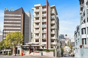 A multi-story building with balconies, a parking area, and a clear sky.