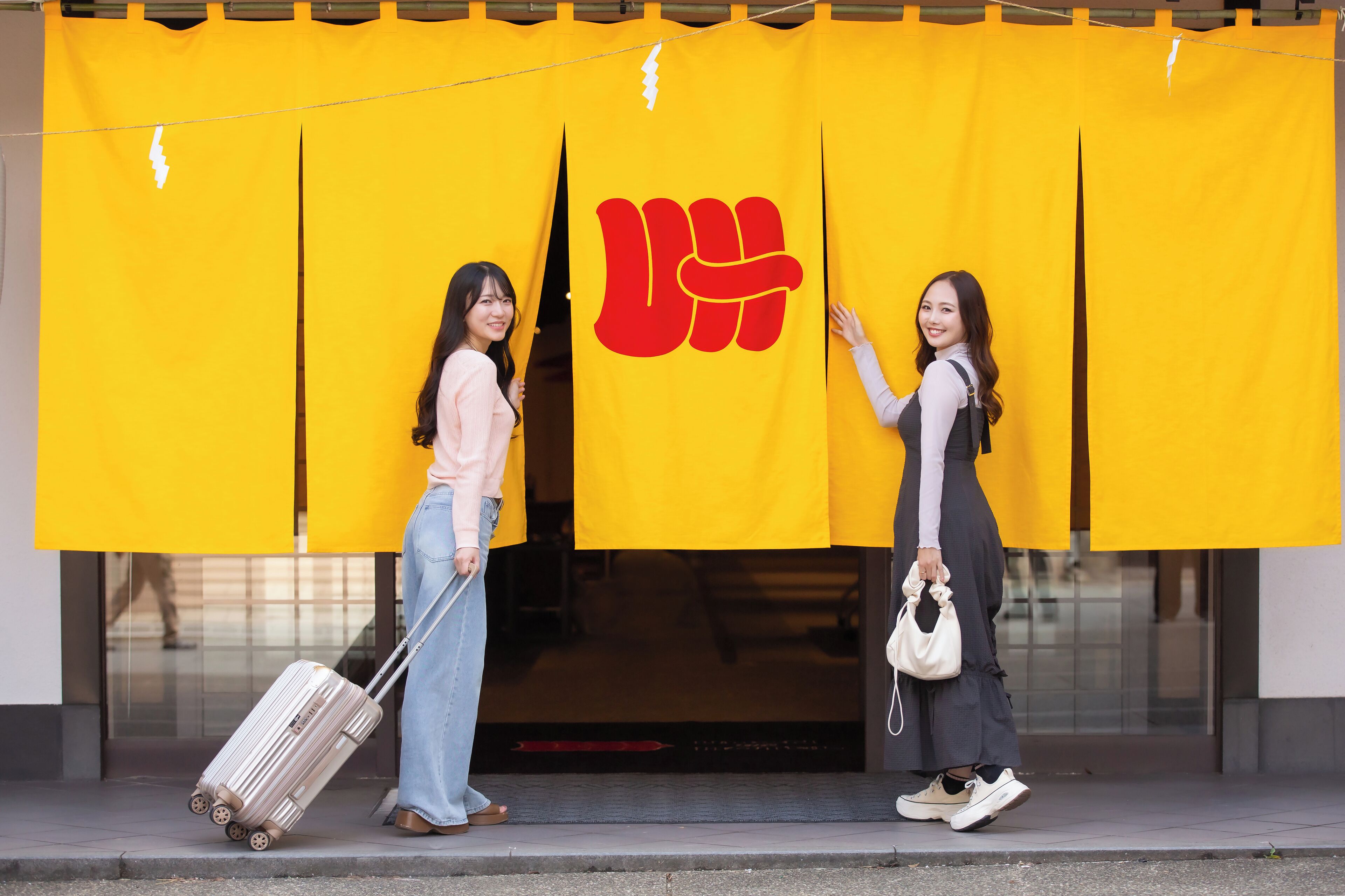 Two individuals standing in front of a yellow curtain with a red symbol, one holding a suitcase and the other a handbag.