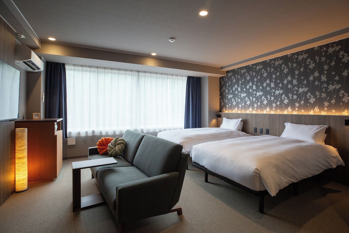 Ein Hotelzimmer mit zwei Betten, einem Schlafsofa, einem Schreibtisch und einem großen Fenster mit Vorhängen.