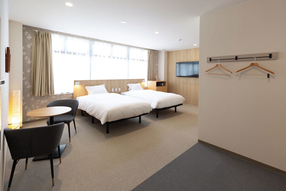 Ein Hotelzimmer mit zwei Betten, einem kleinen runden Tisch und einem Sessel. Zwei an der Wand montierte Fernsehbildschirme und ein Kleiderständer mit Kleiderbügeln sind ebenfalls vorhanden.