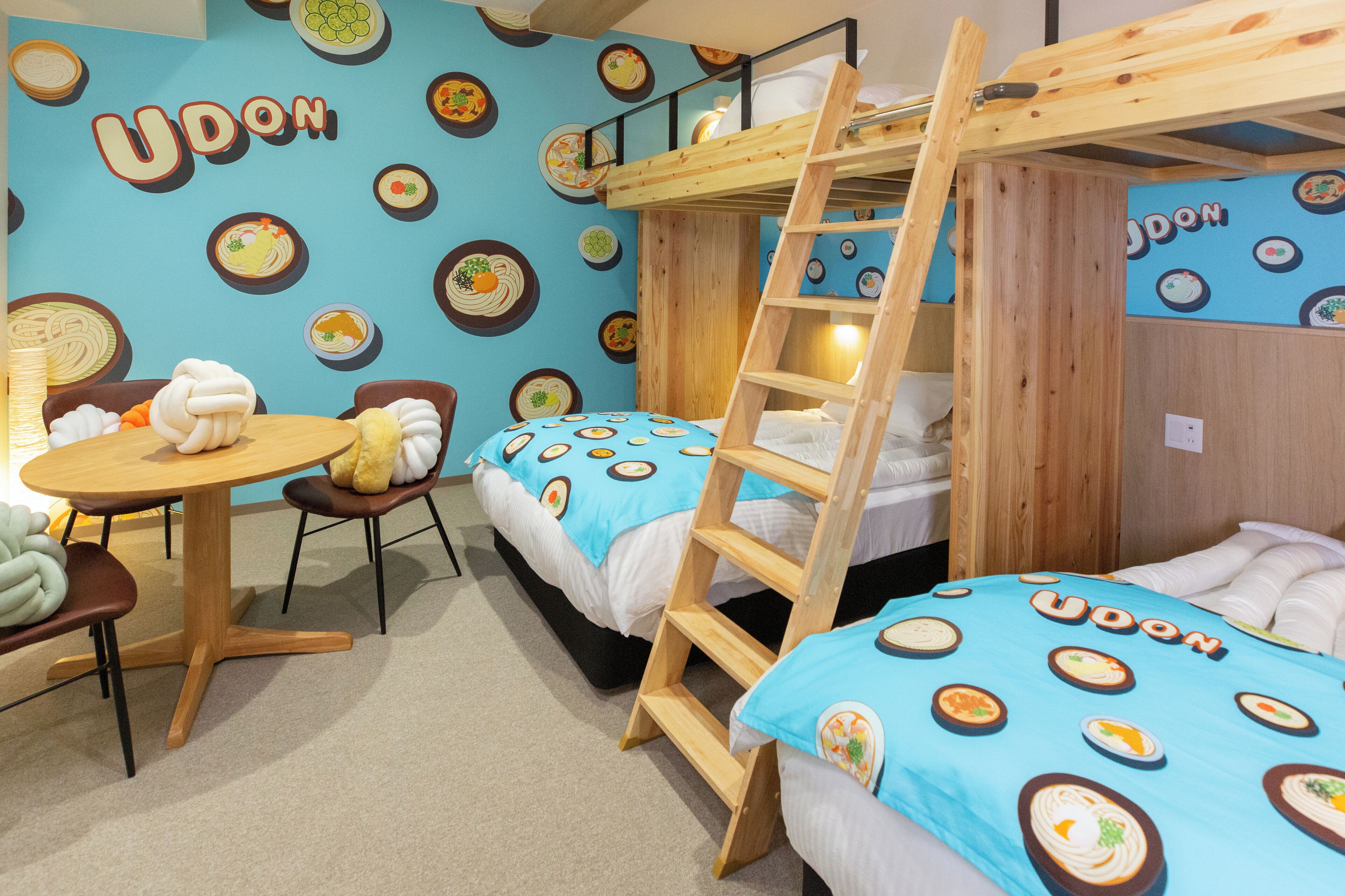 A room with bunk beds, a round table, and chairs, all decorated with a blue tablecloth featuring food patterns.
