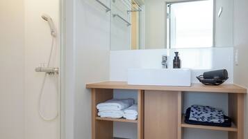 A modern bathroom with a shower, a sink, and a mirror. There are towels and a bag on the shelves.