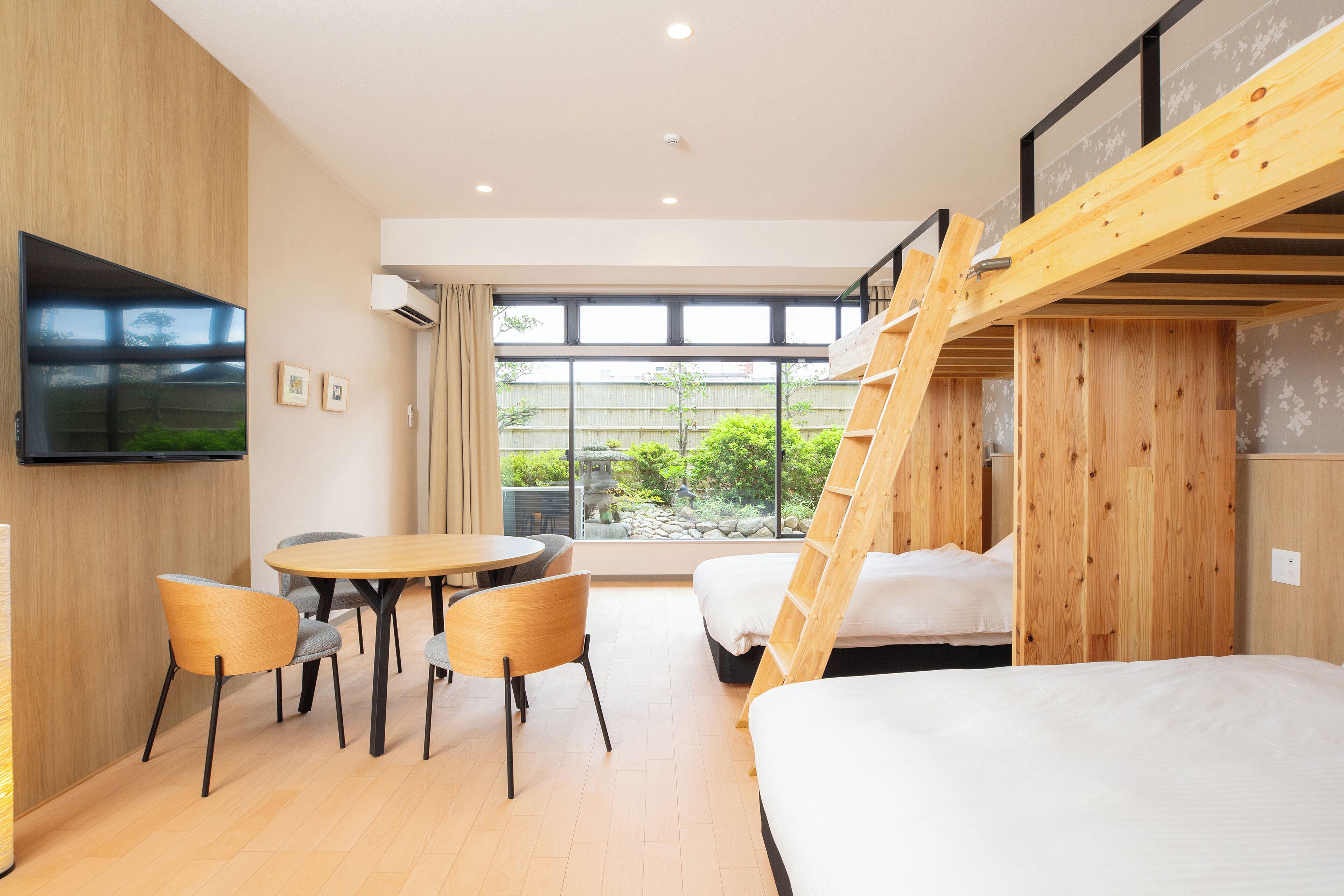 Une chambre d’hôtel moderne avec un lit superposé en bois, un coin repas avec une table et des chaises, un téléviseur à écran plat fixé au mur et de grandes fenêtres offrant une vue sur la verdure environnante.
