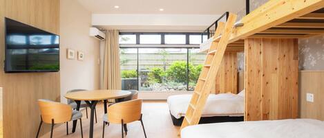 A modern hotel room with a wooden bunk bed, a dining area with a table and chairs, a flat-screen TV mounted on the wall, and large windows offering a view of greenery outside.