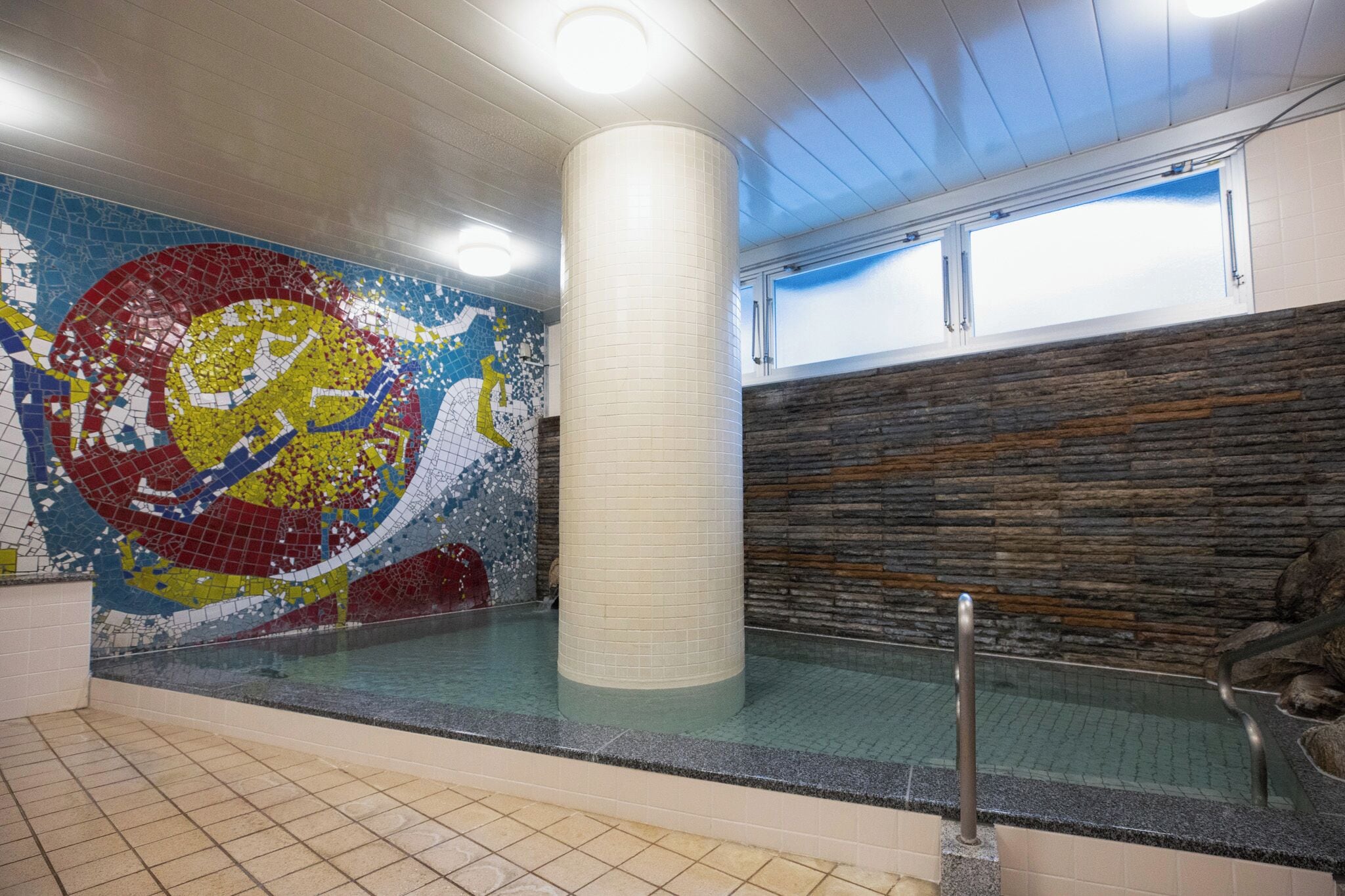 A swimming pool with mosaic artwork on the wall, a stone wall, and a window with a view.