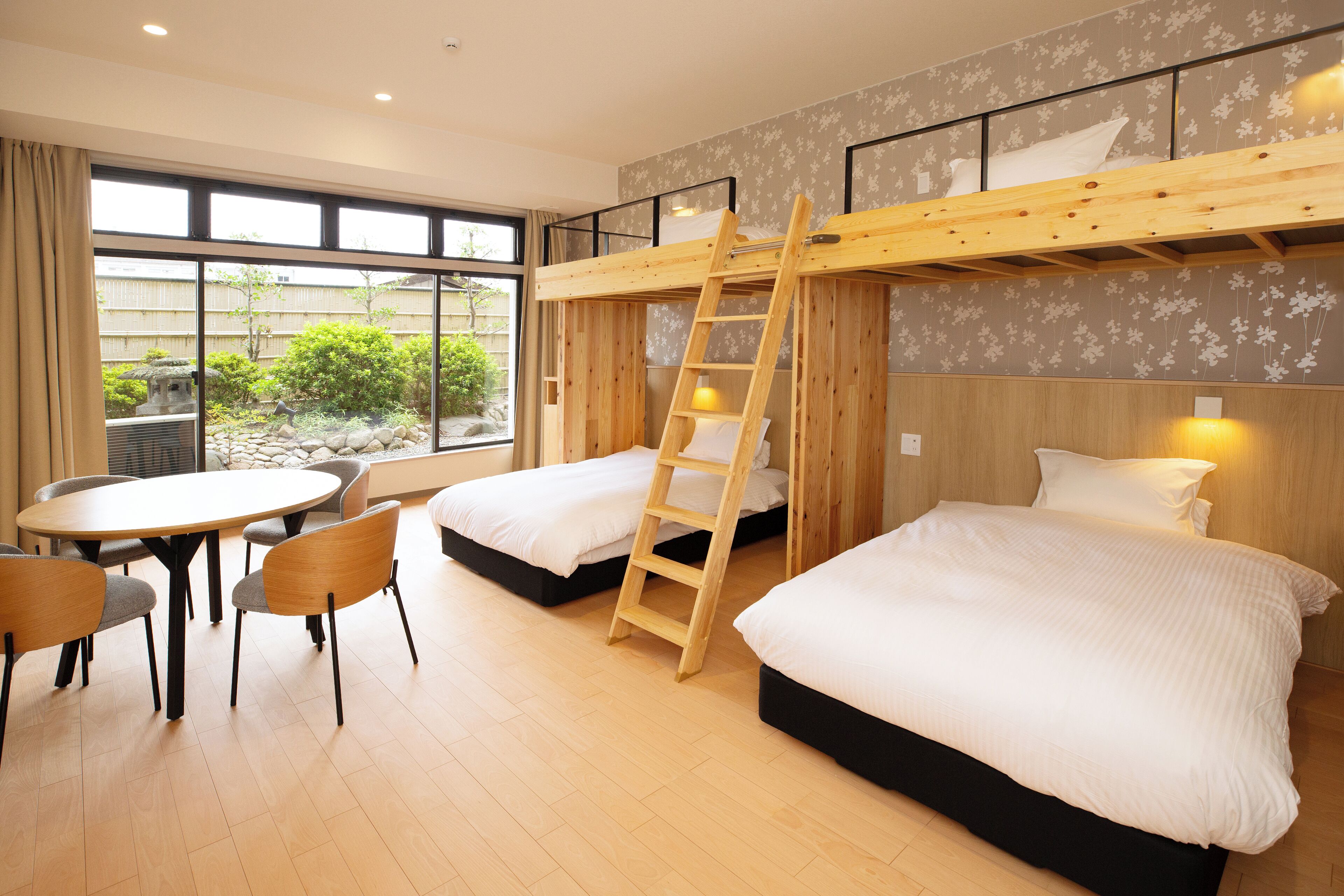 A room with bunk beds, a dining table, chairs, and a view of a garden through large windows.