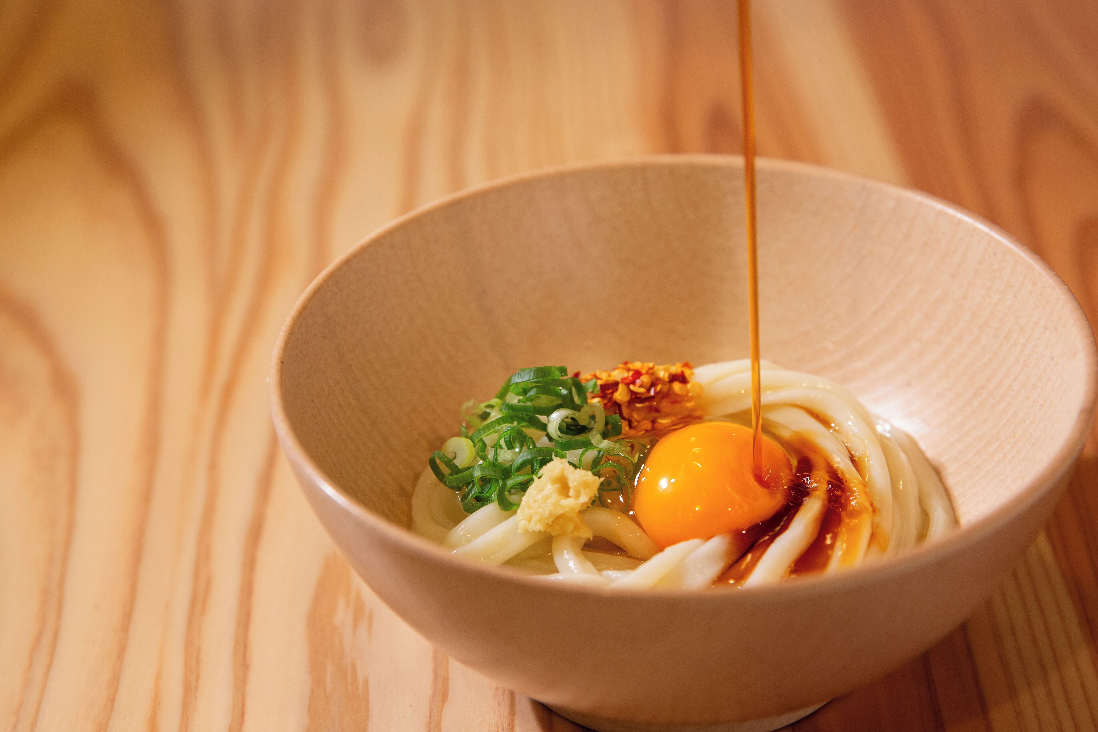 A bowl of ramen with noodles, egg, green onions, and a drizzle of sauce on a wooden table.