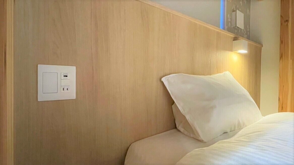 A hotel room with a wooden headboard, a bed with white bedding, and a wall-mounted power outlet.