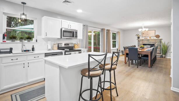 Una cocina moderna con armarios blancos, una isla central con taburetes de bar, un comedor con mesa y sillas de madera, y una chimenea de piedra.
