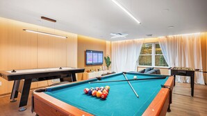 Una mesa de billar con bolas, una mesa de fútbol de mesa y una pantalla de televisión en una habitación con paredes de madera y ventanas amplias.