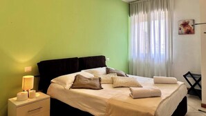 A bedroom with a bed, a nightstand with a lamp, a chair, and a window with curtains. - To complete (Bergamo)
