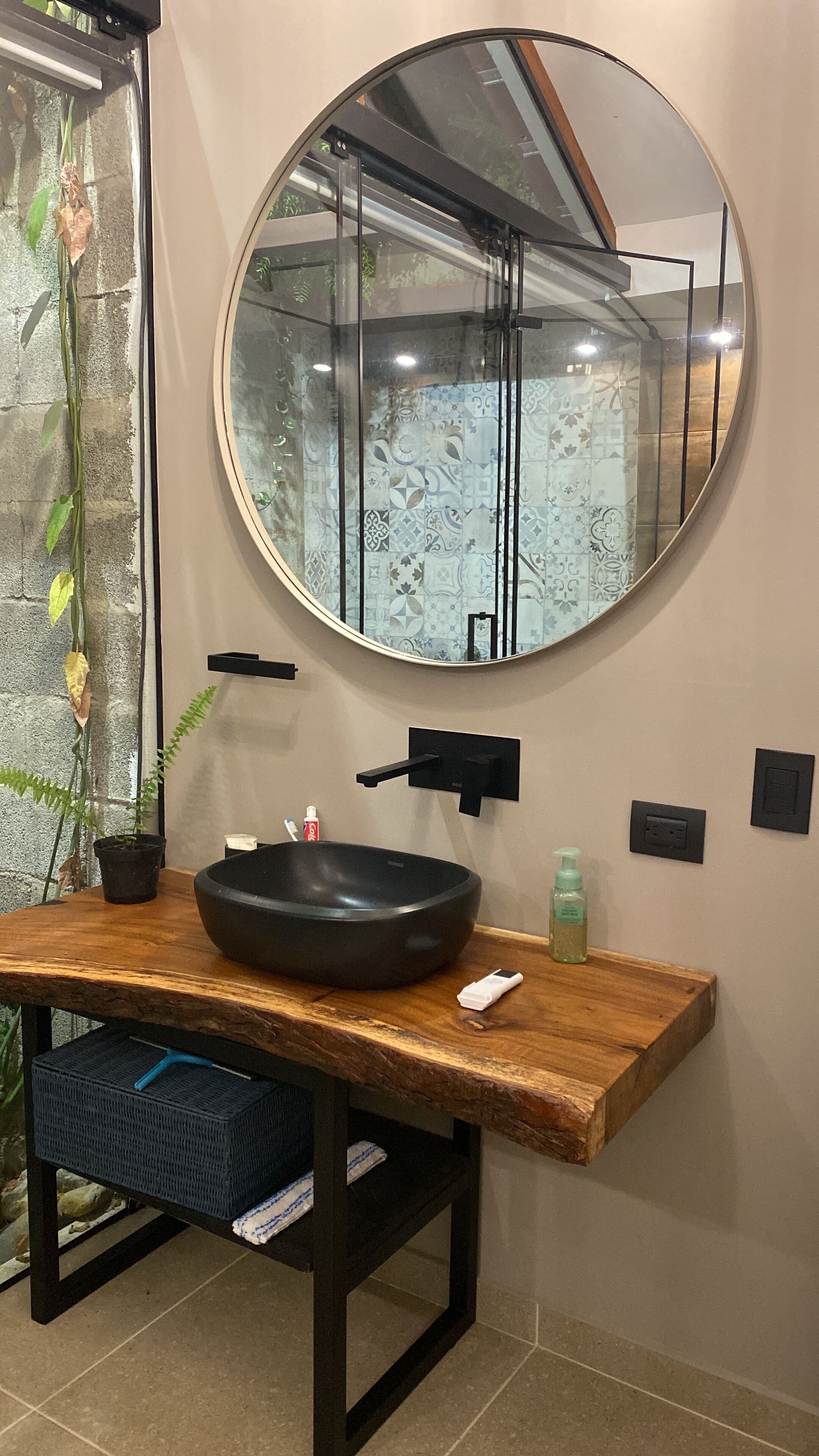 Un baño con un espejo redondo, un lavabo de diseño negro, un mueble de tocador de madera y una planta en maceta.