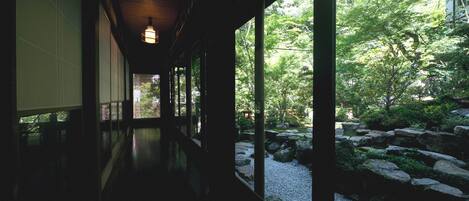 A corridor with wooden flooring and large windows, leading to a garden with rocks and greenery.