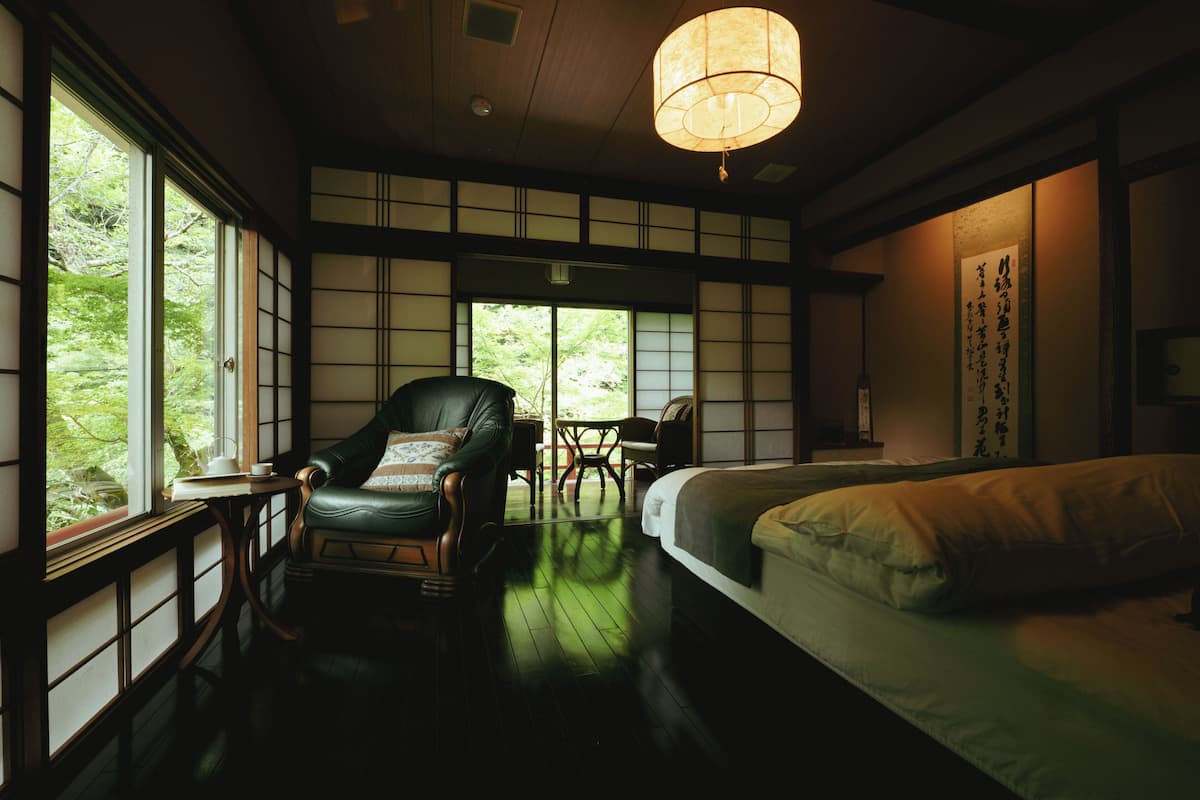 A traditional Japanese-style room with tatami flooring, a low wooden ceiling, a green leather chair, a bed with white bedding, and a large window with a view of greenery.