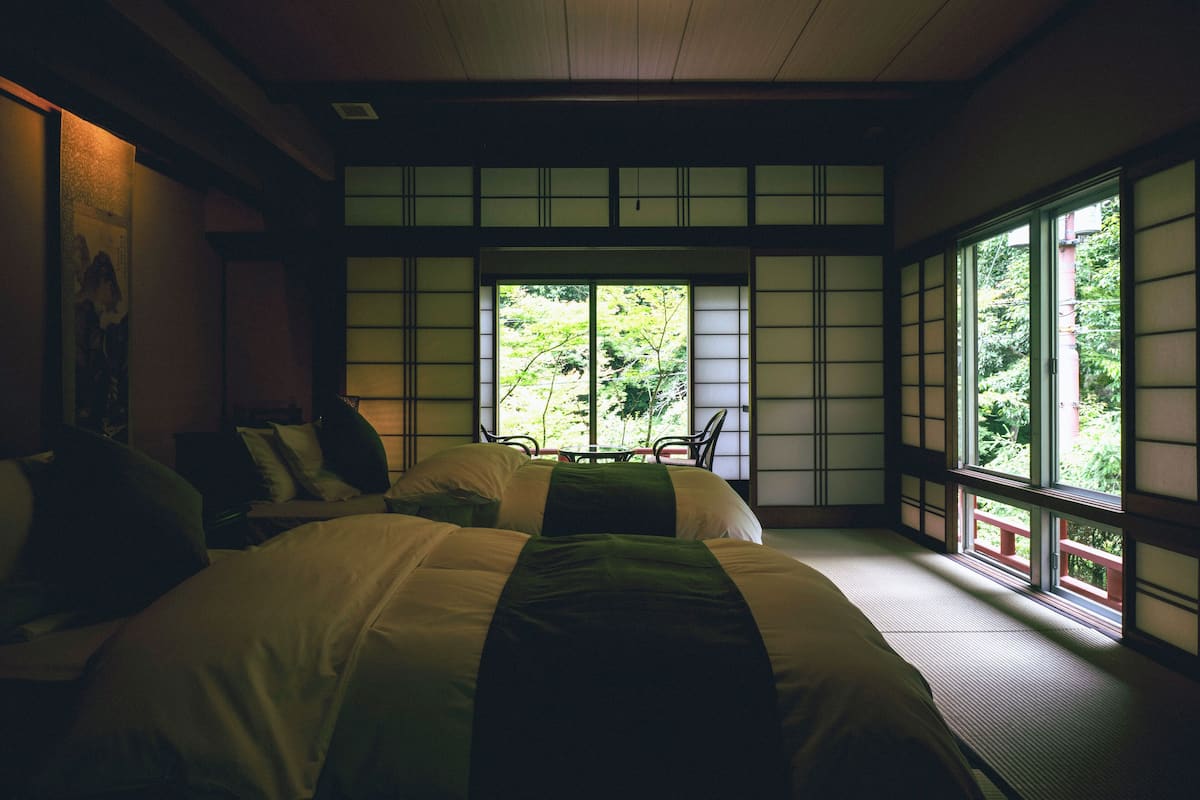 A traditional Japanese-style room with tatami flooring, a low table with a cushion, and a large window offering a view of greenery.