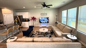 Ein modernes Wohnzimmer mit einer Couch, einem Couchtisch und einem Fernseher. Es gibt eine Küchenzeile mit einem Kühlschrank und Barhocker.
