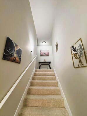Un escalier recouvert de moquette, doté d’une rampe, mène à un palier où se trouvent une petite table et un tableau encadré.