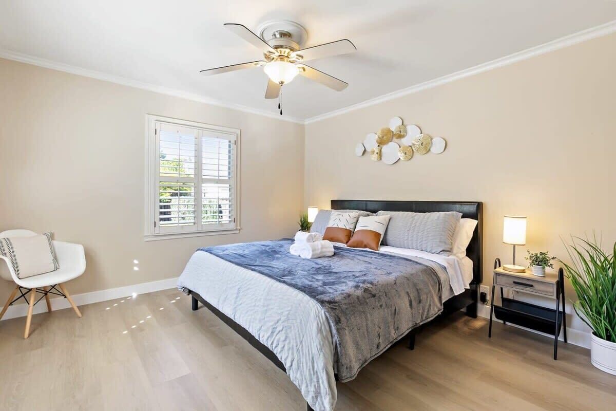 A bedroom with a bed, a ceiling fan, a nightstand with a lamp, a chair, and a window with blinds.