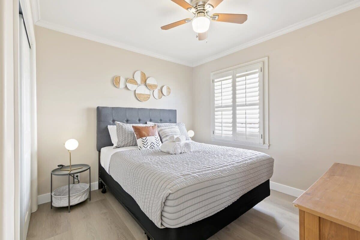 A bedroom with a bed, a ceiling fan, a window with blinds, and a bedside table with a lamp and a basket.