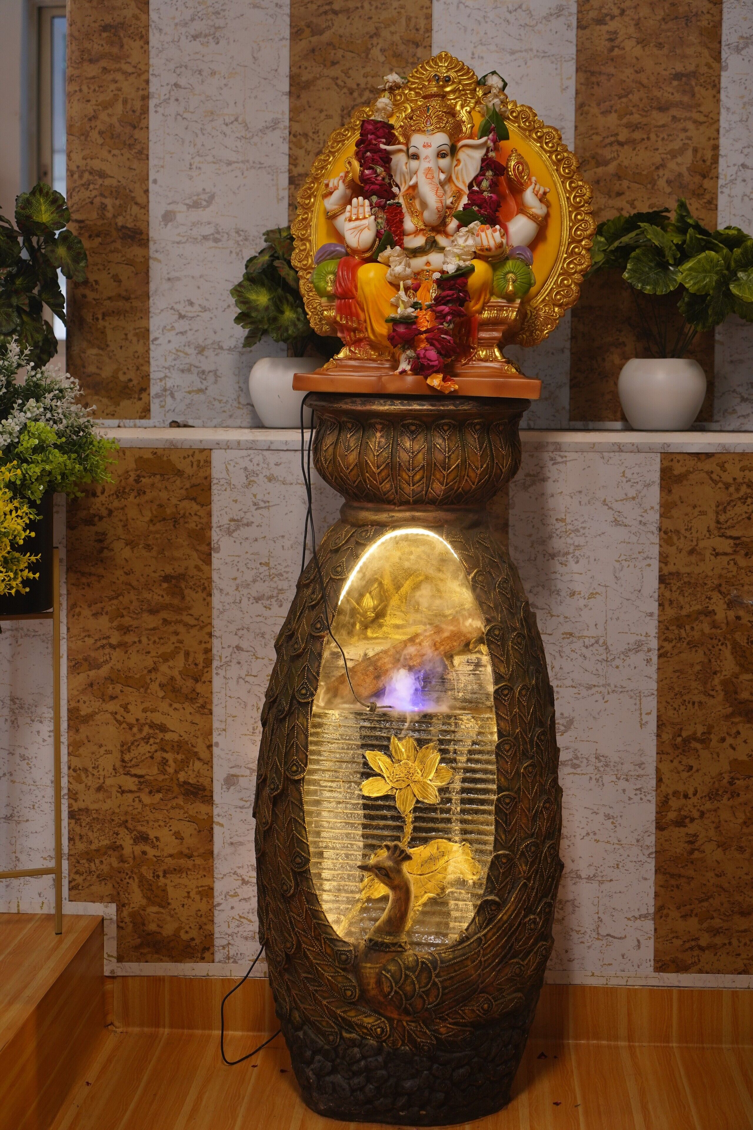 A statue of a deity on a pedestal with intricate designs, placed in a room with a marble wall and potted plants.