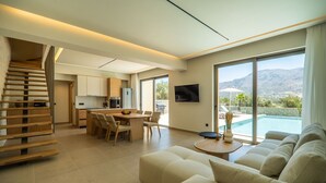 A modern living room with a sofa, a dining area, and a kitchen. There is a pool visible through the large windows. - Villa Zen Chrysi – Vacation villa in Crete (Georgioupolis)