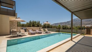 A swimming pool with lounge chairs and umbrellas, surrounded by a stone wall and a view of mountains. - Villa Zen Chrysi – Vacation villa in Crete (Georgioupolis)