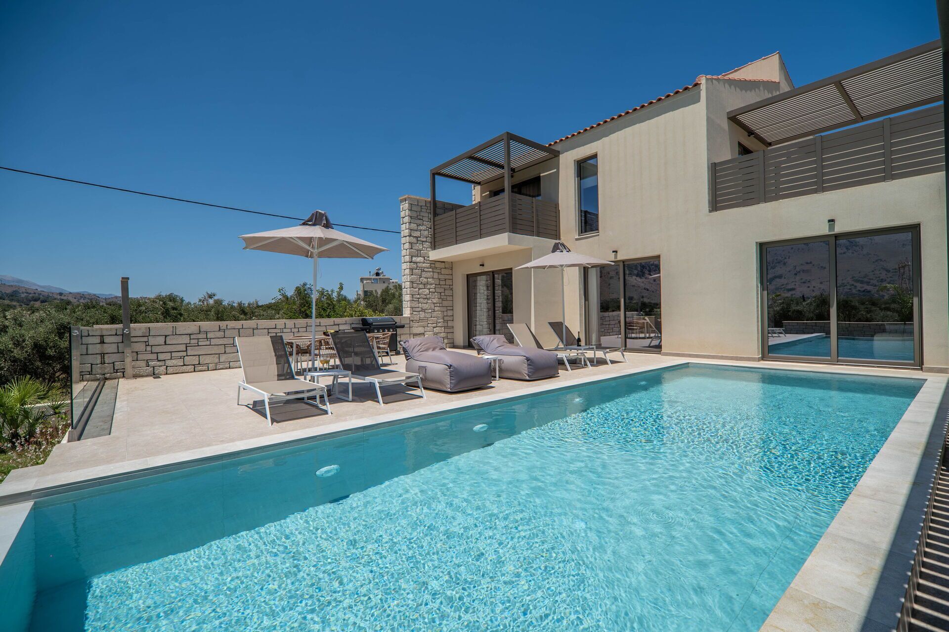A modern poolside area with lounge chairs and umbrellas, a stone wall, and a clear view of the mountains in the background.