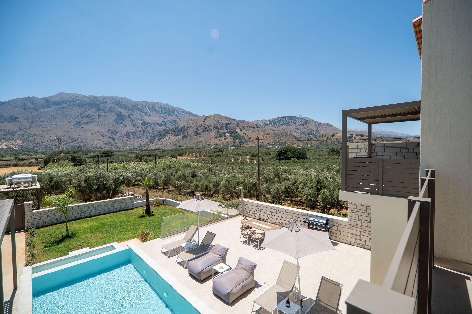 A pool area with lounge chairs, a barbecue, and a view of mountains.