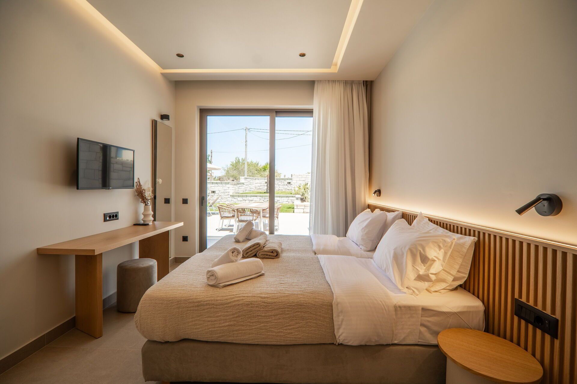 A modern hotel room with a bed, bedside table, TV, and a view of the outdoors.