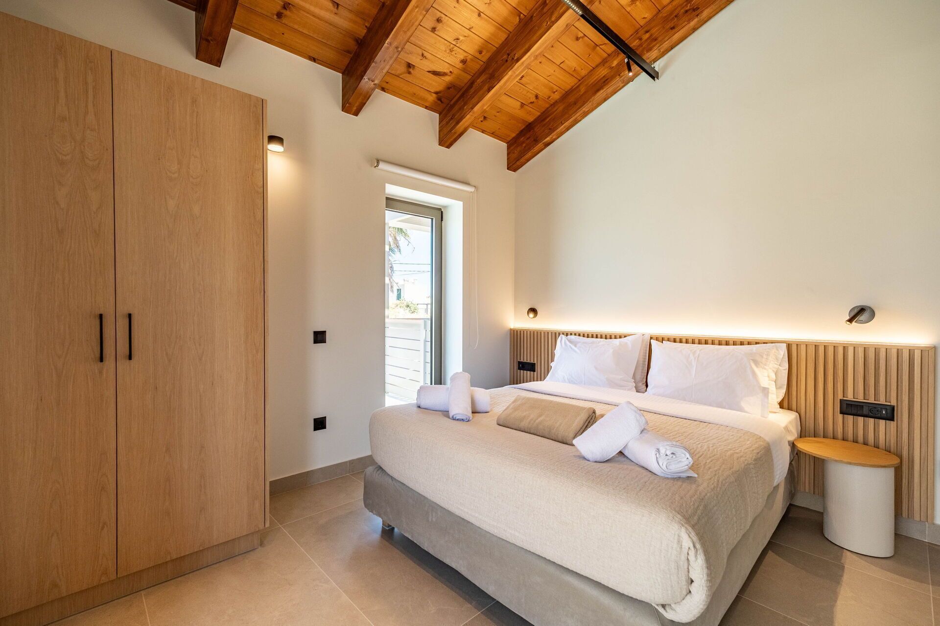A bedroom with a wooden wardrobe, a bed with white linens, a bedside table, and a nightstand.