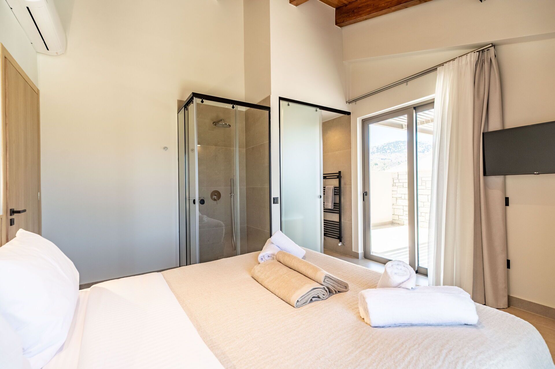 A hotel room with a bed, a glass shower enclosure, a window with curtains, and a flat-screen TV mounted on the wall.