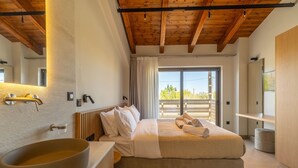 A modern bedroom with a large bed, a wooden ceiling, a large window, and a bathroom with a large basin. - Villa Zen Hera – Vacation villa in Crete (Georgioupolis)