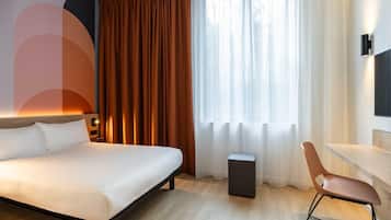 A modern hotel room with a large bed, a desk, a chair, a small stool, and a window with curtains.