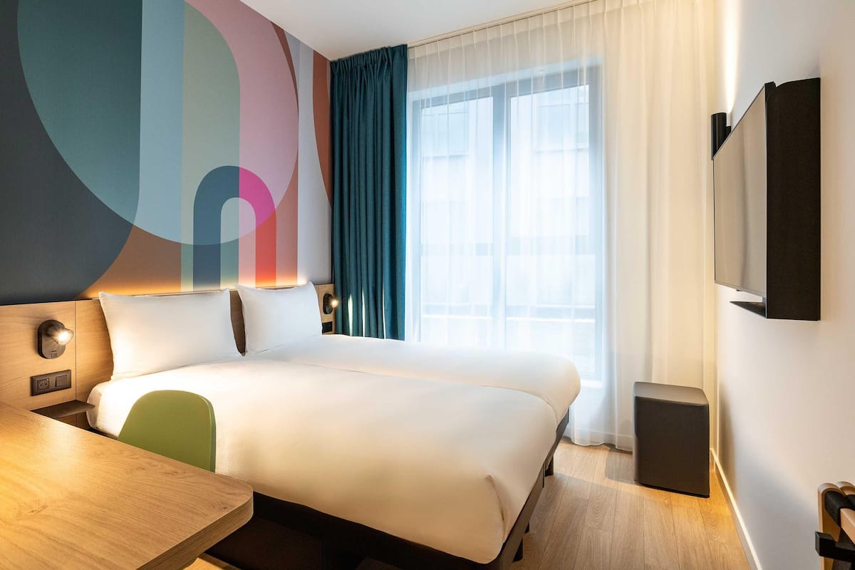A hotel room with a bed, a desk, a chair, a TV, and a window with curtains.