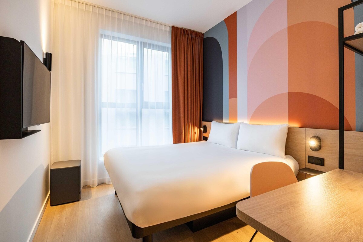 Una habitación de hotel con cama, escritorio, televisión, mesita de noche y ventana con cortinas.