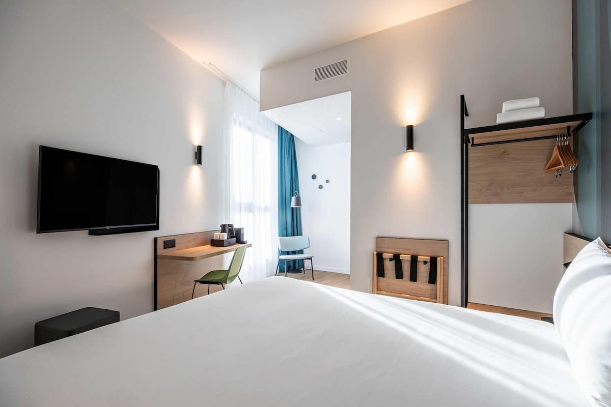 A modern hotel room with a bed, a flat-screen TV, a desk with a chair, a wooden headboard, and a wall-mounted TV.