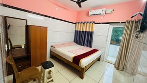 Una camera da letto con un letto, un armadio in legno, una sedia, uno sgabello, una ventola a soffitto, un condizionatore d'aria e una finestra con tende.