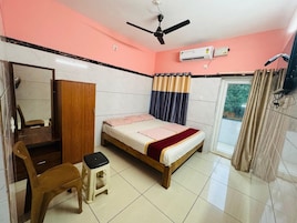 Un dormitorio con cama, armario de madera, silla, taburete pequeño, ventilador de techo, aire acondicionado y ventana con cortinas.