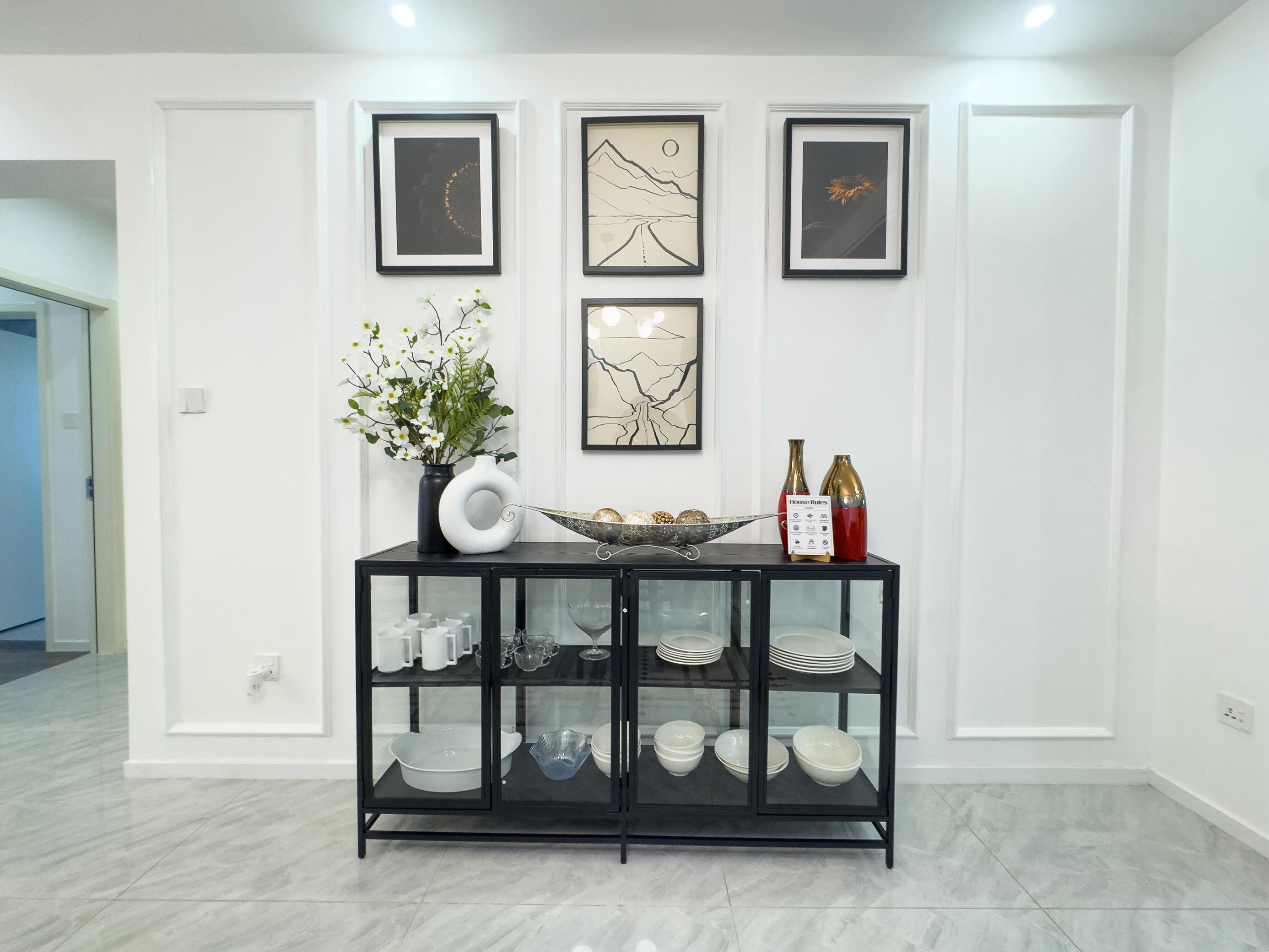 A modern interior with a black glass cabinet displaying white dishes, a vase with white flowers, and three framed artworks on the wall.