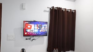 A room with a flat-screen TV mounted on the wall displaying various streaming services, a wooden table with a kettle, remote controls, and bottled water. - Anandam Grand (Greater Noida)